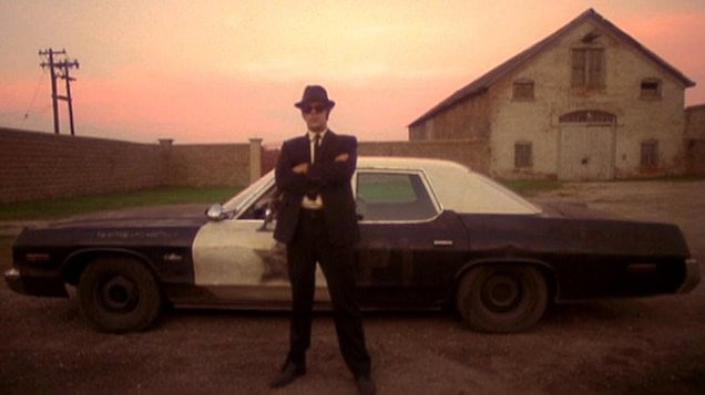 A character from "The Blues Brothers" standing with arms crossed in front of a black and white car.