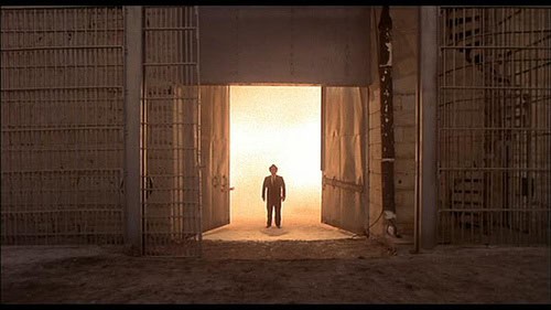 A character from "The Blues Brothers" standing in front of a large open jail door with bright light shining through.