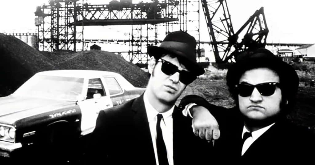 Jake and Elwood Blues from "The Blues Brothers" posing with the Bluesmobile in a black and white photo.